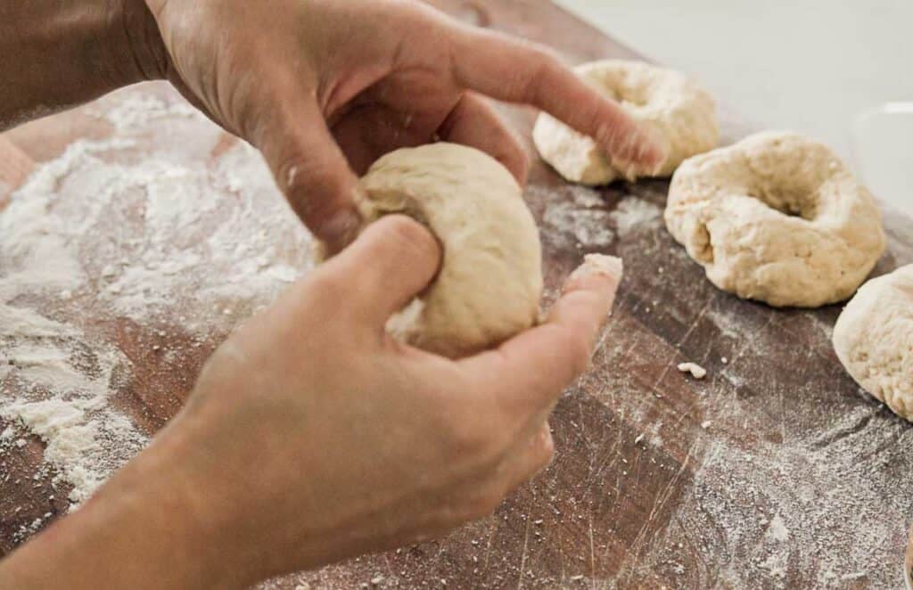 Hands shaping dough into bagels on a floured wooden surface, with three other unbaked bagels in the background.